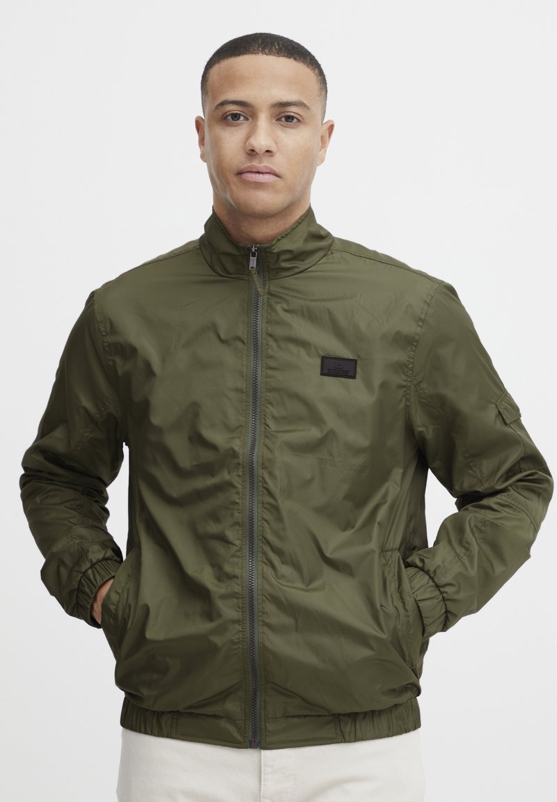 Olive green zippered jacket with a stand-up collar, elastic cuffs, side pockets, and a small black logo patch on the chest. Smooth, lightweight fabric.
