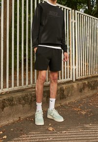 Person klädd i svart Nike-tröja och shorts med vita Nike-strumpor och ljusgröna Nike-sneakers som står vid ett vitt metallstaket.