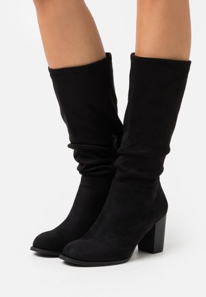 Paire de bottes noires mi-mollet à talons carrés portées sur des jambes sur un fond blanc uni.
