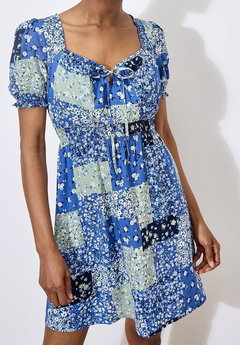 Femme portant une robe bleue à patchwork floral avec des manches bouffantes et un col noué sur fond blanc.