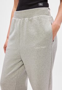 Szare dresy z elastycznym pasem, bocznymi kieszeniami i białym logo "Calvin Klein" na dolnej części nogawki. Gładki materiał bawełniany.