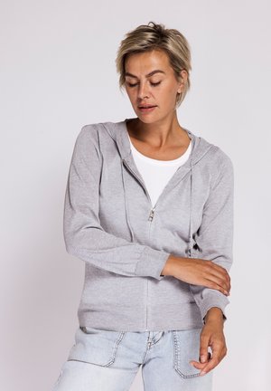 Frau mit hellgrauem Zip-Hoodie, weißem T-Shirt und hellblauen Jeans, die ihren Ärmel vor einem einfarbigen hellen Hintergrund zurechtrückt.