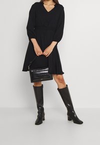 Svart klänning med långa ärmar, V-ringning och smockad midja, kombinerad med knähöga svarta läderstövlar och en strukturerad svart handväska.