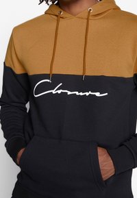 Felpa a blocchi di colore in cotone marrone e nero, con tasca frontale, polsini a coste e cappuccio con cordini. Dettaglio del logo bianco sul petto.