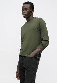 Jauns vīrietis stāv ar vienu roku kabatā, tērpies olīvzaļā garajā piedurknē un melnās biksēs pret vienkāršu gaišu fona.