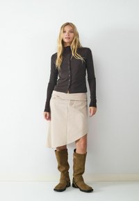 Braun langärmliges Hemd mit Knöpfen, beiger asymmetrischer Rock und knielange braune Wildlederstiefel mit Schnallen. Schlichte, strukturierte Stoffe.