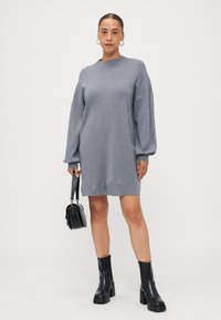 Gray knitted sweater dress with dropped shoulders, long sleeves, and a relaxed fit, paired with black heeled ankle boots and a small black handbag.