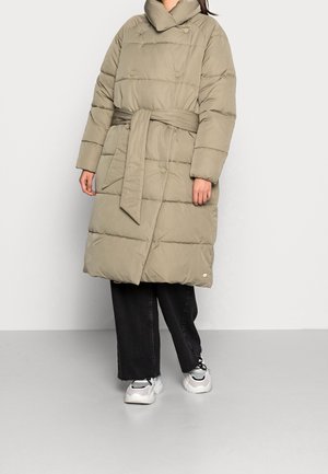 Cappotto lungo imbottito in verde oliva con colletto alto, vita cinturata, grandi bottoni e texture a coste. Abbinato a pantaloni neri a gamba larga e sneakers.