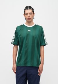 Mlada osoba s ispletenom kosom, odjevena u zelenu Adidas majicu s bijelim prugama na rukavima i tamnoplavim hlačama, na jednostavnoj pozadini.