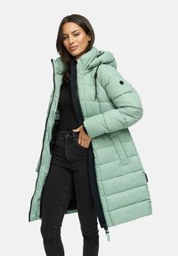 Kvinna iklädd en lång ljusgrön vadderad vinterjacka med huva, svart topp och högmidjade svarta jeans mot en vit bakgrund.