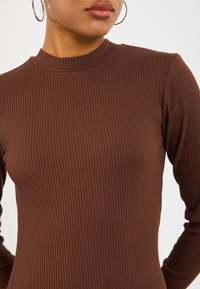 Donna che indossa grandi orecchini a cerchio e un top lungo a maniche ribbed marrone con colletto a lupetto, mostrata dalle spalle alle labbra.
