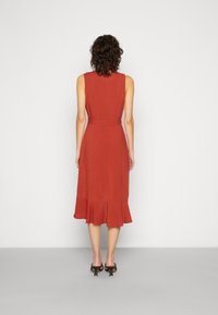 Vestido sem mangas de cor ferrugem com corpete ajustado, cintura cingida e bainha com volantes, feito de tecido leve. Usado com sapatos de salto pretos.