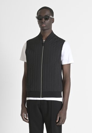 Gilet imbottito nero con collo alto, zip frontale e orlo a costine, indossato sopra una maglietta bianca. Modello con occhiali da sole scuri.