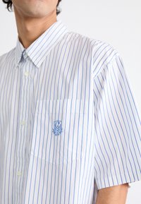 Valka puuvillainen, lyhythihainen kauluspaita, jossa on vaaleansiniset pystysuorat raidat. Paidassa on rintatasku sekä taskuun brodeerattu logo.