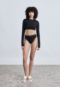 Μαύρο crop top με μακριά μανίκια και μαγιό σλιπ με ψηλή μέση, διακοσμημένα με λευκή λωρίδα με αντίθετο λογότυπο. Λευκά σαγιονάρες. Λείο ύφασμα, εφαρμοστή γραμμή.