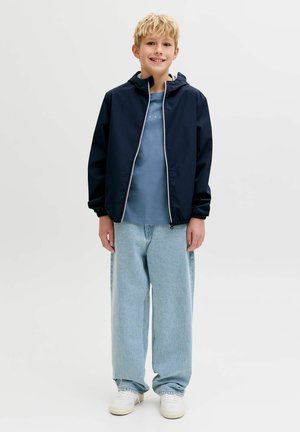 Garçon blond souriant, portant une veste zippée bleu marine, un t-shirt bleu, un jean ample bleu clair et des baskets blanches, debout sur un fond uni.