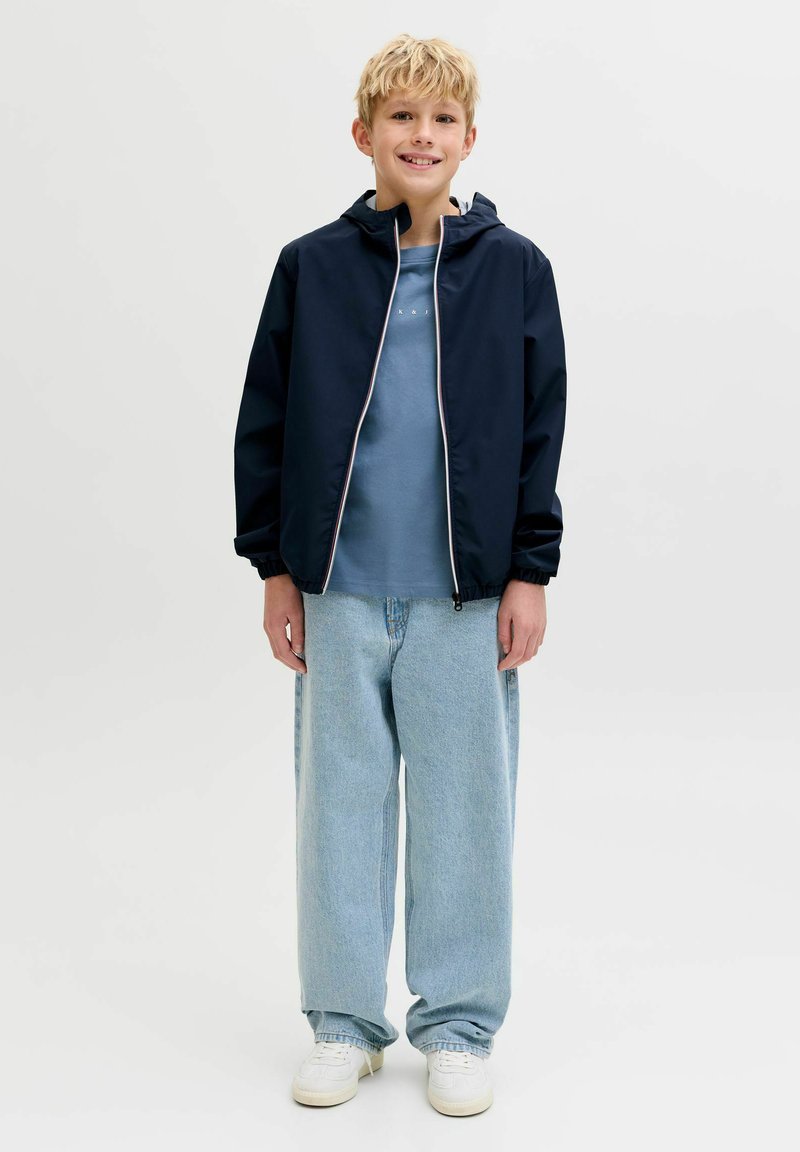 Lyshåret dreng smilende, iført en marineblå lynlåstrøje, blå t-shirt, løse lyseblå jeans og hvide sneakers, stående på en ensfarvet baggrund.