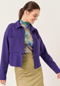 Paarse cropped jas met twee voorkruikezakken en knoopsluiting, gelaagd over een veelkleurige, gestructureerde top met hoge hals, gecombineerd met een beige rok.