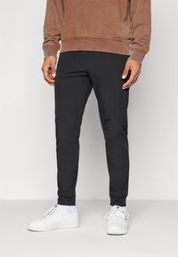 Pantaloni sportivi neri con vestibilità affusolata, vita elasticizzata e tasche con zip, abbinati a sneaker bianche e un felpa marrone.