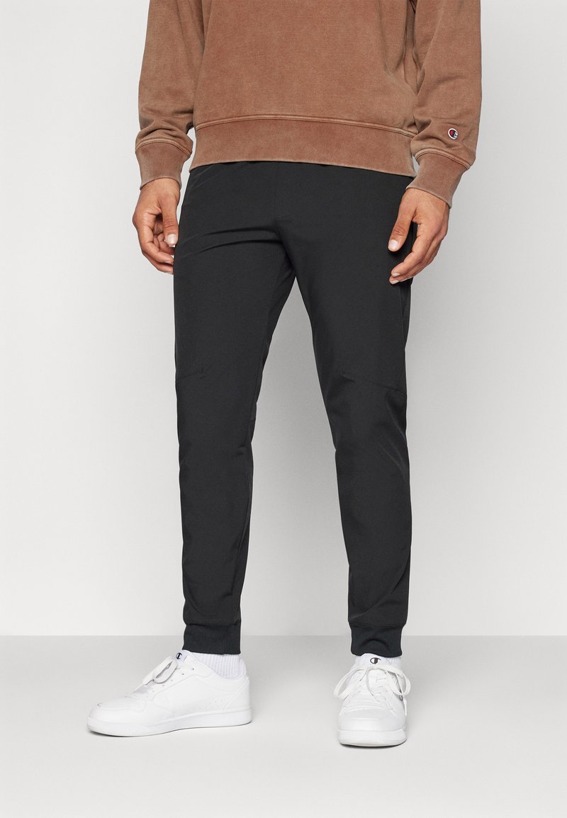Pantaloni sportivi neri con vestibilità affusolata, vita elasticizzata e tasche con zip, abbinati a sneaker bianche e un felpa marrone.