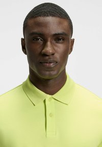 Kragepolo i ljus limegrön, tillverkad av mjukt tyg med en texturerad krage och placket med två knappar, som visar en figursydd design.