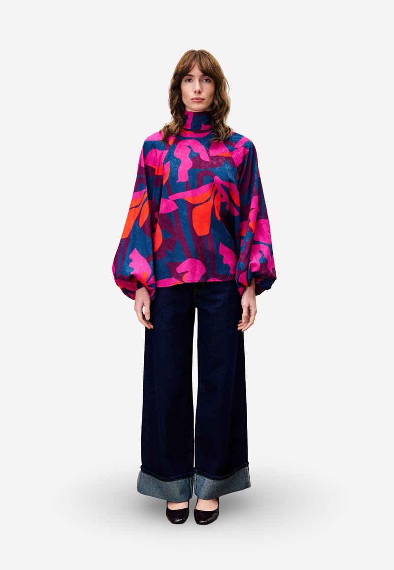 Haut à imprimé floral violet et rose avec un col montant et des manches bouffantes, associé à un jean en denim foncé à jambes larges avec un ourlet contrasté.