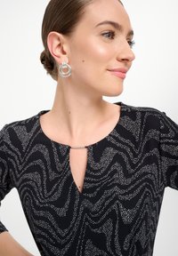 Zwart met patronen jurk met witte abstracte golven, met een sleutelgat decolleté en metalen accent. Model draagt zilveren creolen.