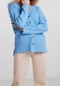 Ljusblå ribbad cardigan med knappstängning, V-ringning och vågig nederkant, i kombination med beiga raka byxor. Mjuk textur, avslappnad passform.