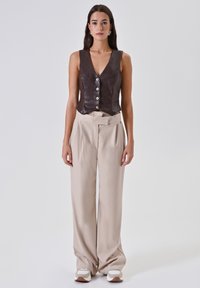 Gilet in pelle marrone con cinque bottoni bianchi, abbinato a pantaloni beige a vita alta e sneakers bianche, su uno sfondo neutro.