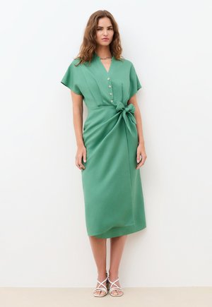 Frau trägt ein grünes Midikleid mit Knöpfen und einer seitlichen Schleife, steht vor einem schlichten weißen Hintergrund und trägt weiße Riemchensandalen.