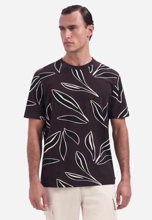 Man die een zwart T-shirt met korte mouwen draagt met grote witte bladpatronen, gecombineerd met lichtbeige broek, staand tegen een effen achtergrond.
