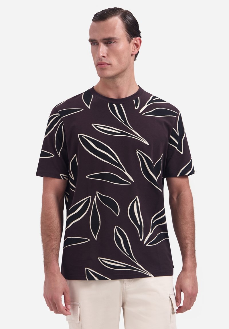 Man die een zwart T-shirt met korte mouwen draagt met grote witte bladpatronen, gecombineerd met lichtbeige broek, staand tegen een effen achtergrond.