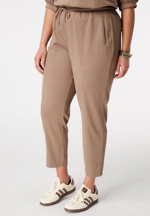 Personne portant un pantalon texturé marron clair à taille élastique et des baskets crème avec des rayures marron, debout sur un fond blanc.