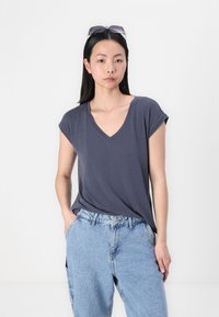 Γκρι T-shirt με V λαιμόκοψη και κοντά μανίκια, συνδυασμένο με ανοιχτό μπλε τζιν παντελόνι. Η μαλακή υφή και η χαλαρή γραμμή είναι χαρακτηριστικά στοιχεία.
