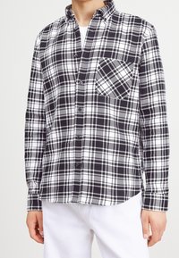 Camicia a quadri bianca e nera con colletto a punta, taschino frontale e maniche lunghe, realizzata in un tessuto liscio con un motivo a scacchi.