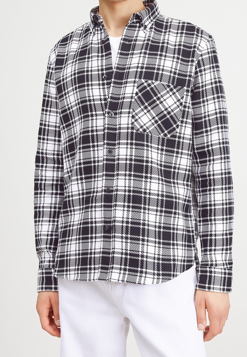 Camicia a quadri bianca e nera con colletto a punta, taschino frontale e maniche lunghe, realizzata in un tessuto liscio con un motivo a scacchi.