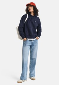 Navy hoodie with front pocket, white drawstrings, paired with light blue wide-leg jeans and a red cap. Cotton fabric, casual design.