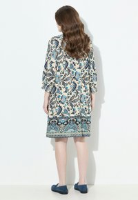 Mujer con largo cabello castaño ondulado lleva un vestido beige con patrones de flores y paisley azules, combinado con zapatos planos azules, de pie contra una pared blanca.