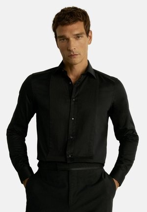 Mees kannab musta pika varrukaga särki volditud esipaneeliga ja musti pükse, käed taskutes, neutraalne ilme, valge taust.