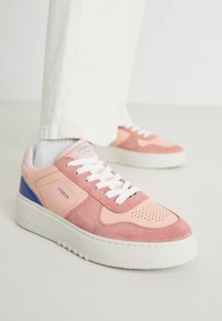 Baskets roses et corail avec des matériaux en daim et en cuir, des lacets blancs et une semelle en caoutchouc texturée. Présente des accents bleus sur le talon.