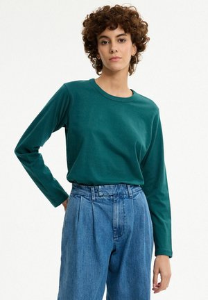 Mujer con cabello rizado y corto que lleva una camisa de manga larga verde oscuro y vaqueros azules de talle alto, de pie frente a un fondo blanco.