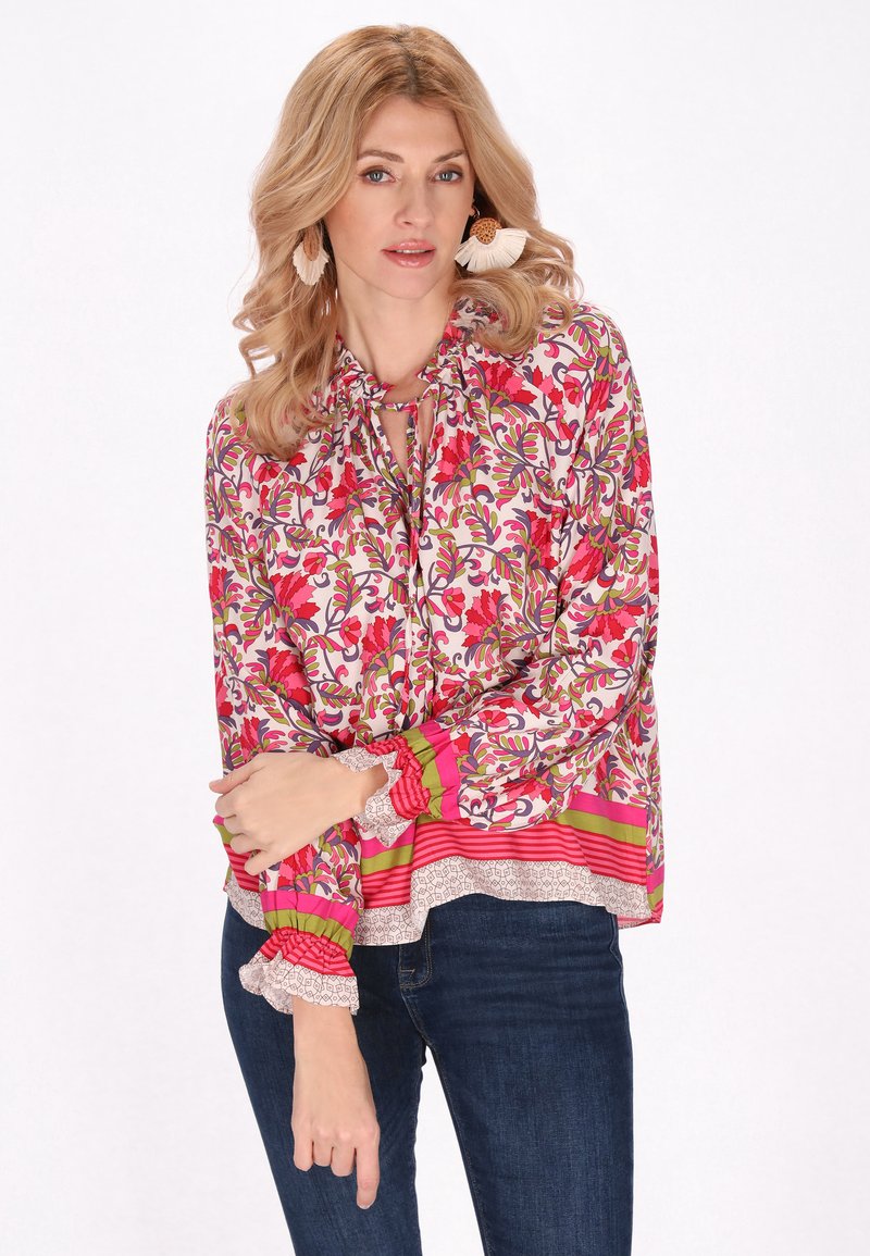 Blomsterbluse med snorelukning ved halsen, prydet med et miks af pink, grønne og hvide mønstre samt rynkede manchetter. Båret med mørke jeans.