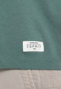 Vihreä puuvillapaita, jossa on valkoinen etiketti, jossa lukee "ESPRIT" ja "Established 1968" -teksti, sekä tekstuurillinen ompeluyksityiskohta helman ympärillä.