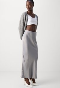Fustă maxi din satin gri deschis, cu o textură netedă, cusături laterale, o siluetă fluidă, purtată cu un top alb scurt și un cardigan tricotat gri.