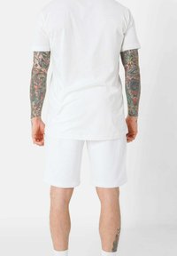 Set di t-shirt in cotone bianco e pantaloni corti con una vestibilità ampia. La t-shirt ha maniche corte; i pantaloni corti sono lunghi fino al ginocchio. Tatuaggi visibili sulle braccia.