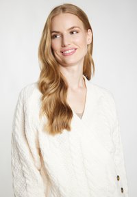 Witte kabelgebreide cardigan met een wikkelontwerp, voorzien van grote knopen en textuurdetails. Zacht, dik gebreid materiaal, elegante val.
