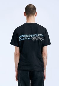 Černé bavlněné tričko s krátkým rukávem, na zadní straně s modrým textem: "DRDENIM OD ROKU 2004 (Dobré časy)" s grafickými detaily.