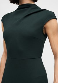 Κοντινό πλάνο ενός ατόμου που φοράει αμάνικο σκουροπράσινο φόρεμα με ψηλό λαιμό και εφαρμοστή μέση, πάνω σε απλό φόντο.