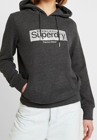 Dunkelgrauer Hoodie mit einer Fronttasche, der ein silbernes Logo und Text zeigt. Mit Kordelzug an der Kapuze sowie gerippten Bündchen und Saum.