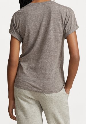 Tummaihoinen henkilö, joka pukeutunut harmaaseen t-paitaan ja vaaleanharmaisiin housuihin, seisoo takanaan kameraan päin, toinen käsi taskussa.